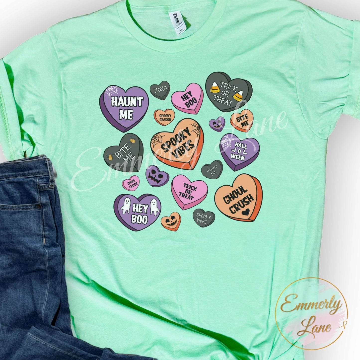 A green unisex t-shirt with a Halloween themed design featuring various playful phrases and icons related to Halloween, positioned next to a pair of blue jeans.