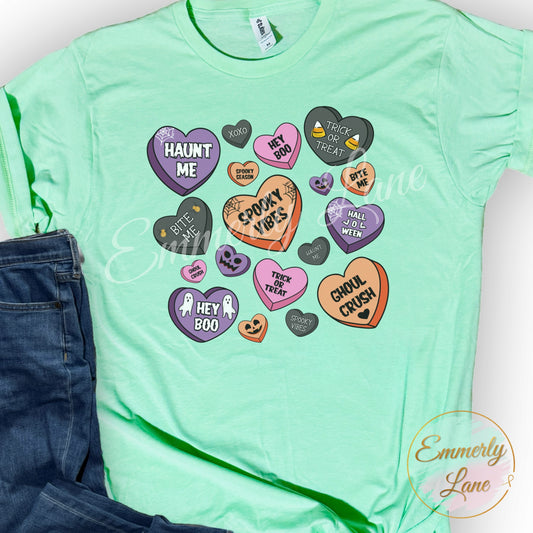 A green unisex t-shirt with a Halloween themed design featuring various playful phrases and icons related to Halloween, positioned next to a pair of blue jeans.