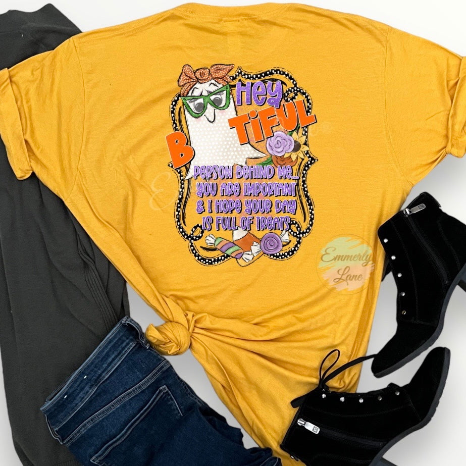 A heather mellow yellow t-shirt showing the back of the shirt with graphic print of a creamy colored ghost with wearing green glasses and an orange bow. The image says Hey Beautiful (with the ghost replacing the letters "e-a-u") person behind me...you are important & I hope your day is full of treats. The flat lay picture also displays black heeled boots,blue jeans and a cardigan. 