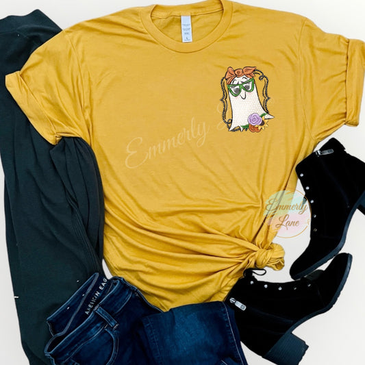 A yellow t-shirt with a graphic print of a creamy colored ghost with white dots wearing an orange bow and floral flowers on the bottom of the ghost. Displayed flat with a pair of black shoes, cardigan and blue jeans.